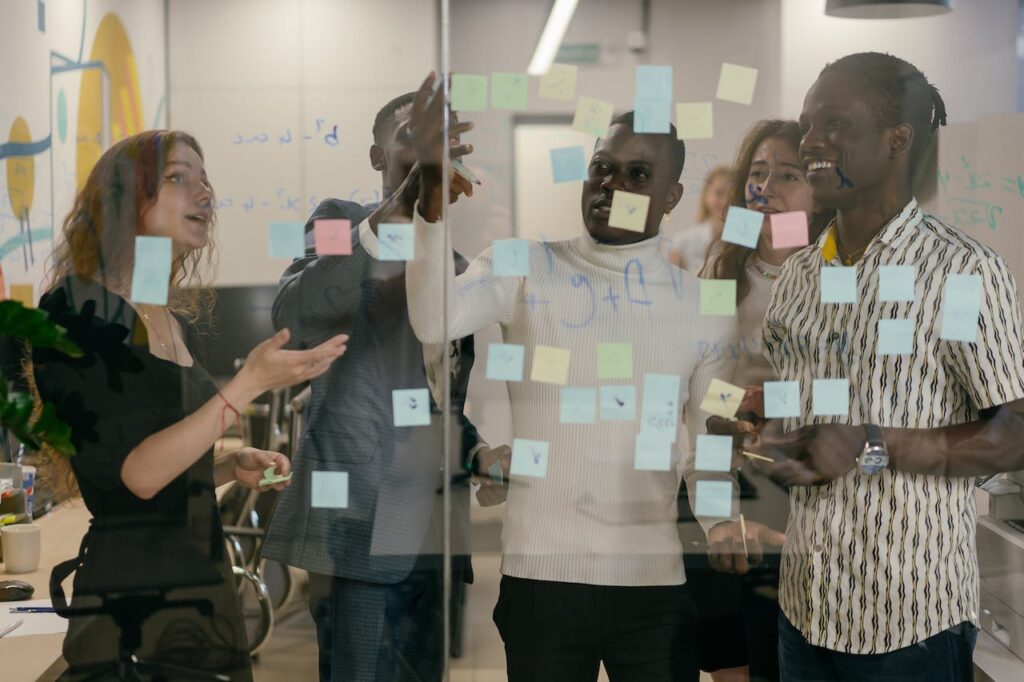 Five diverse professionals collaborating in a modern office, with the Facilitator on the left leading the discussion. The group stands around a glass wall covered with colorful sticky notes, actively brainstorming and sharing ideas. Photo by Mikhail Nilov: https://www.pexels.com/photo/colleagues-looking-at-sticky-notes-9301872/